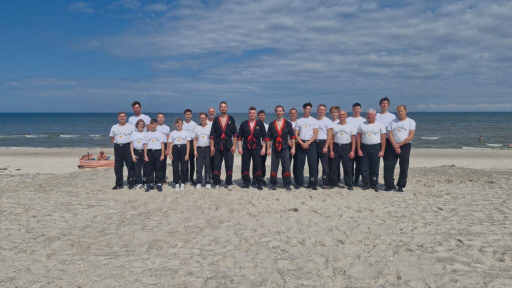 Grupa uczniów Wing Tsun w jednolitych strojach pozuje na plaży nad morzem
