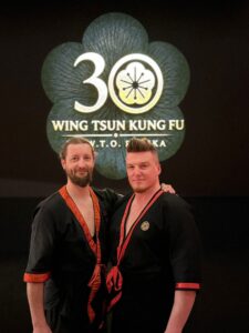 Dwie osoby w strojach Wing Tsun pozują przed ciemną planszą z logo Wing Tsun Kung Fu i liczbą 30