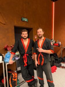 Dwóch mężczyzn w czarnych strojach Wing Tsun z czerwonym wykończeniem pozuje za kulisami