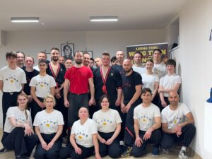 Duża grupa uczniów i instruktorów Wing Tsun w białych koszulkach i czarnych strojach pozuje w sali
