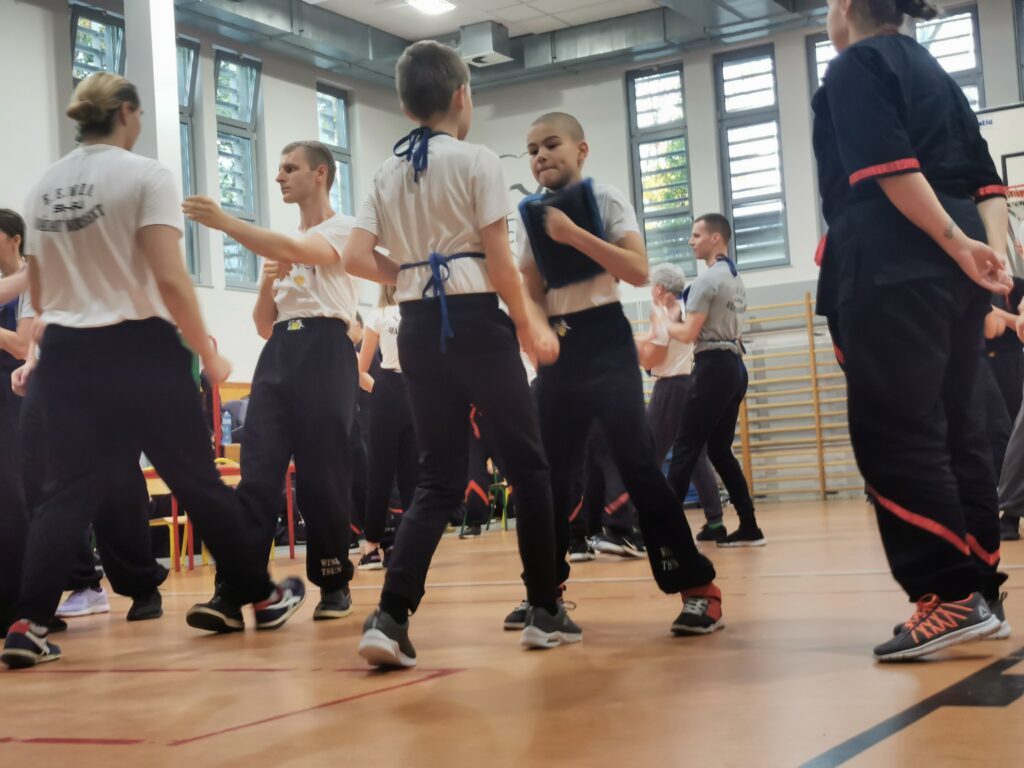 Dzieci i dorośli ćwiczą w parach w sali, trener pokazuje technikę uczniom