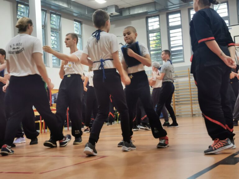 Dzieci i dorośli ćwiczą w parach w sali, trener pokazuje technikę uczniom